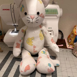 May include: A handmade stuffed cat toy crafted from white fabric featuring a repeating pattern of yellow bunnies, pink clouds, and blue beach balls. The cat has black button eyes, a pink embroidered nose, and stitched whiskers on its face.