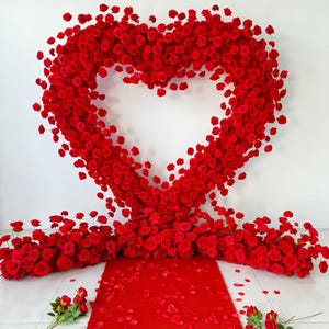 May include: A large heart-shaped arch constructed from numerous vibrant red roses. The arch stands above a red carpet runner, which is flanked by clusters of red roses and scattered red rose petals on the floor, creating a floral display.