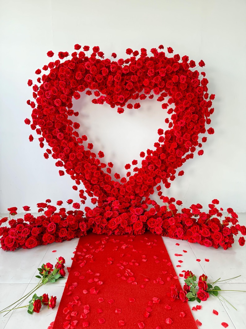 May include: A large heart-shaped arch constructed from numerous vibrant red roses. The arch stands above a red carpet runner, which is flanked by clusters of red roses and scattered red rose petals on the floor, creating a floral display.