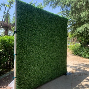 May include: A green artificial hedge backdrop for photography or events. The backdrop is made of a realistic-looking artificial grass and is supported by a metal stand.