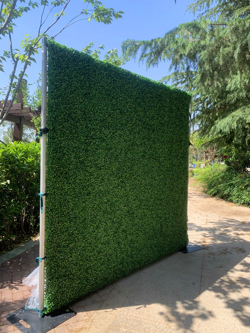 May include: A green artificial hedge backdrop for photography or events. The backdrop is made of a realistic-looking artificial grass and is supported by a metal stand.