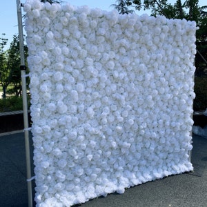 May include: White artificial flower wall backdrop for events, weddings, or photo shoots. The wall is made of many small white roses.