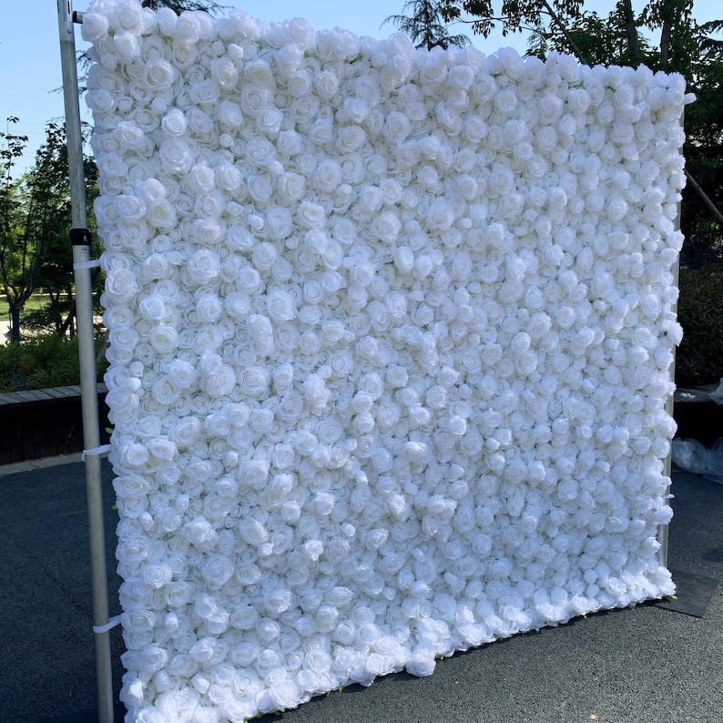 May include: White artificial flower wall backdrop for events, weddings, or photo shoots. The wall is made of many small white roses.
