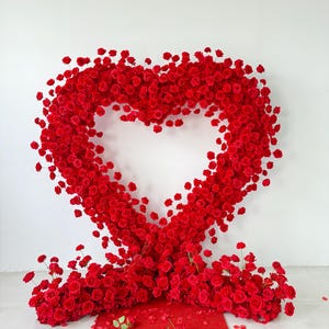 May include: A large heart-shaped floral arrangement constructed from numerous vibrant red roses. The structure features a hollow center and rests on a base of dense red roses, with scattered red rose petals on the floor in front of the display.