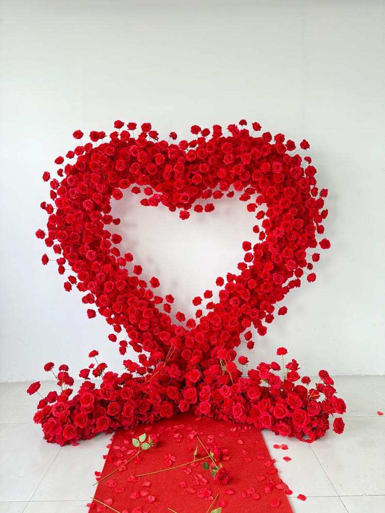 May include: A large heart-shaped floral arrangement constructed from numerous vibrant red roses. The structure features a hollow center and rests on a base of dense red roses, with scattered red rose petals on the floor in front of the display.