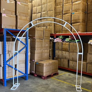 May include: White metal archway standing in a warehouse setting. The archway is made of metal tubing and has a simple, modern design. The archway is standing on a concrete floor in front of a wall of cardboard boxes.