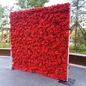 May include: A large red rose flower wall backdrop for events, weddings, or parties. The wall is made of artificial flowers and is attached to a metal frame.