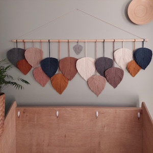 May include: A wall hanging made of macrame with a wooden dowel and a variety of coloured leaves. The leaves are in shades of brown, beige, grey, and blue.