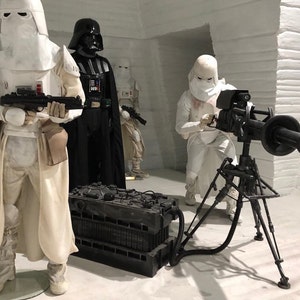 Peut inclure: Un groupe de personnages de Star Wars, dont Dark Vador et des Stormtroopers, se tiennent dans un décor enneigé. Une grosse arme noire est montée sur un trépied.