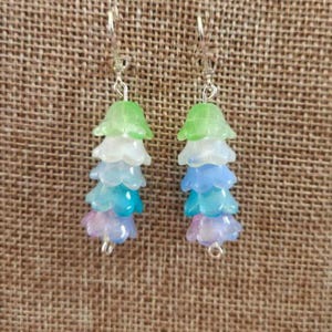 May include: A pair of drop earrings with a layered floral design. Each earring has a silver hook and is made up of translucent petals in green, white, blue, and purple. The earrings are set against a textured, neutral background.