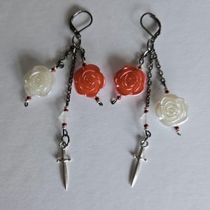 May include: A pair of dangle earrings featuring white and orange rose-shaped beads, small red beads, and silver-colored dagger charms. The earrings are connected by dark metal chains and hooks.