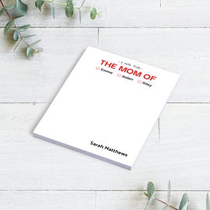 May include: White notepad with the text "The Mom Of" in red. There are three checkboxes below the text, labeled "Emma", "Aiden", and "Riley". The bottom of the notepad says "Sarah Matthews".