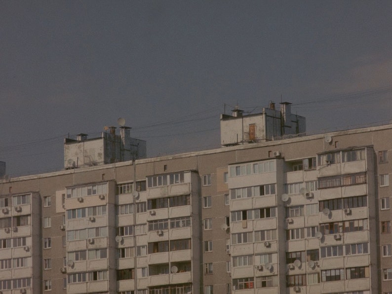 May include: A large, gray apartment building with many windows and balconies. The building has a flat roof and is made of concrete.