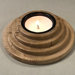 May include: A wooden candle holder with a black metal ring and a lit tea light candle in the center. The holder has a layered design with four concentric circles.