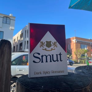 May include: A pack of cigarettes with a white background and a burgundy top. The word "Smut" is printed in large, black letters, with "Dark Spicy Romance" below. The pack is resting on a wooden post.