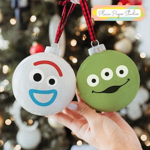 Pu&ograve; includere: Due palline di Natale rotonde, una bianca e una verde, sono tenute contro uno sfondo sfocato di luci natalizie. L'ornamento bianco ha una faccia da cartone animato con un sorriso rosso e una bocca blu. L'ornamento verde ha una faccia da cartone animato con tre occhi e un sorriso nero.