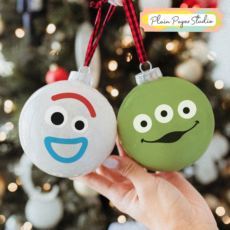 Pu&ograve; includere: Due palline di Natale rotonde, una bianca e una verde, sono tenute contro uno sfondo sfocato di luci natalizie. L'ornamento bianco ha una faccia da cartone animato con un sorriso rosso e una bocca blu. L'ornamento verde ha una faccia da cartone animato con tre occhi e un sorriso nero.