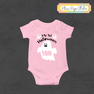 K&ouml;nnte beinhalten: Hellrosa Baby-Body mit Halloween-Motiv. Der Body hat eine wei&szlig;e Geistergrafik mit dem Wort "BOO!" in Rosa und dem Text "My 1st Halloween". Zwei kleine Geistergrafiken befinden sich &uuml;ber dem Text.