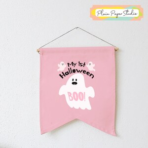K&ouml;nnte beinhalten: Ein rosa Banner mit dem Text "My 1st Halloween" und einer Geisterillustration mit dem Wort "BOO!". Das Banner ist an einer Holzstange mit einer Schnur aufgeh&auml;ngt.