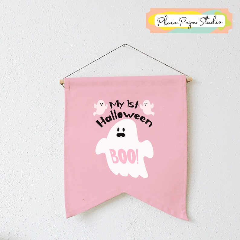 K&ouml;nnte beinhalten: Ein rosa Banner mit dem Text "My 1st Halloween" und einer Geisterillustration mit dem Wort "BOO!". Das Banner ist an einer Holzstange mit einer Schnur aufgeh&auml;ngt.
