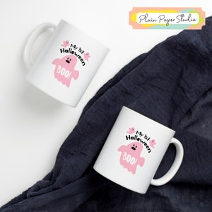 K&ouml;nnte beinhalten: Zwei wei&szlig;e Keramikbecher mit Halloween-Motiv. Jeder Becher zeigt einen rosa Geist mit dem Text "My 1st Halloween BOO!". Die Becher stehen auf einem dunkelblauen Stoffhintergrund.