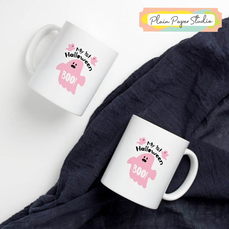 K&ouml;nnte beinhalten: Zwei wei&szlig;e Keramikbecher mit Halloween-Motiv. Jeder Becher zeigt einen rosa Geist mit dem Text "My 1st Halloween BOO!". Die Becher stehen auf einem dunkelblauen Stoffhintergrund.