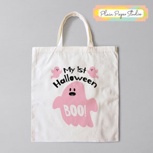 K&ouml;nnte beinhalten: Eine wei&szlig;e Canvas-Tragetasche mit cremefarbenen Griffen. Die Tasche zeigt eine rosa Geistergrafik mit dem Wort "BOO!" und dem Text "My 1st Halloween". Zwei kleine rosa Geistergrafiken sind dar&uuml;ber.
