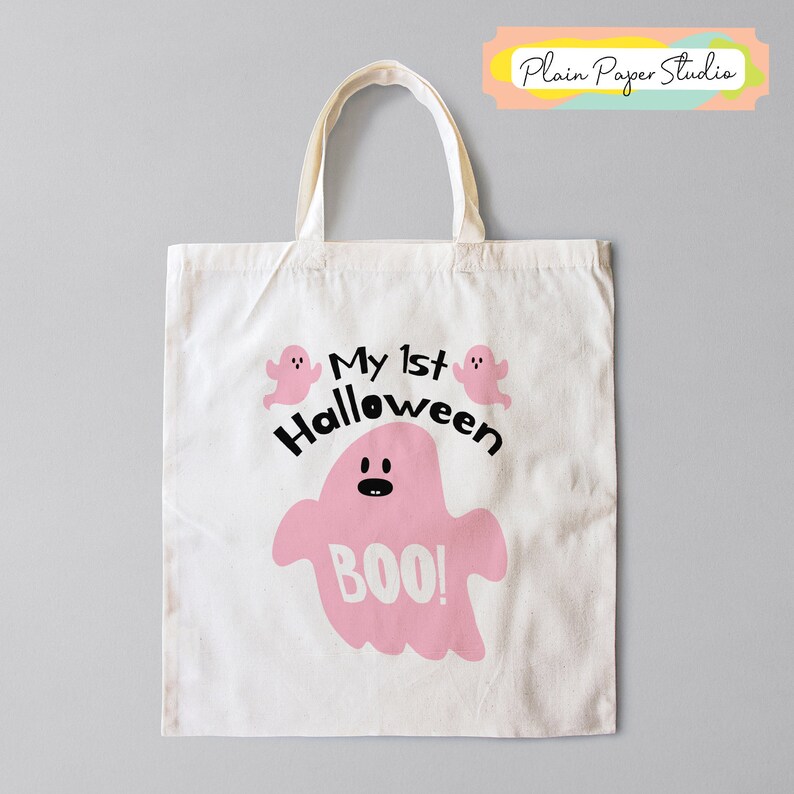 K&ouml;nnte beinhalten: Eine wei&szlig;e Canvas-Tragetasche mit cremefarbenen Griffen. Die Tasche zeigt eine rosa Geistergrafik mit dem Wort "BOO!" und dem Text "My 1st Halloween". Zwei kleine rosa Geistergrafiken sind dar&uuml;ber.