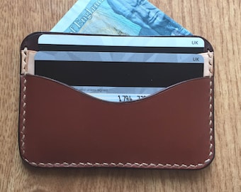 May include: A brown leather wallet with a stitched edge and a flap that folds over. The wallet has two cards inside and a bill sticking out of the top.