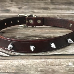 Könnte beinhalten: Ein braunes Lederhundekragen mit silbernen Metallspikes. Das Halsband liegt auf einer Holzoberfläche.