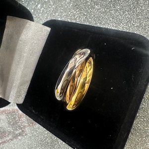 May include: A three-band ring with a silver, gold, and rose gold finish, displayed in a black velvet jewelry box. The bands are interlocked, creating a stylish and modern design. The ring is a piece of jewelry.