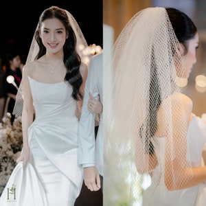 May include: A bride wearing a white wedding dress with a long veil. The veil is a fine mesh with a delicate pattern. The bride is walking down the aisle with her arm linked with a person in a white suit.