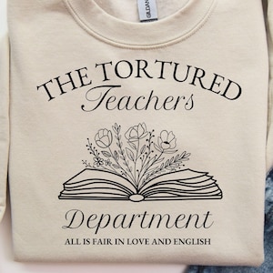 May include: A beige sweatshirt with the text "THE TORTURED Teachers Department" and the phrase "ALL IS FAIR IN LOVE AND ENGLISH". The design includes an open book with floral illustrations.