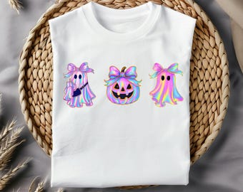 May include: White t-shirt featuring three Halloween-themed designs. The designs include two ghosts and a pumpkin, all adorned with pastel bows and a holographic, iridescent effect. One ghost holds a drink and a small purse. The shirt is displayed on a woven basket.