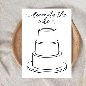 Puede incluir: Una tarjeta blanca con el texto "decorate the cake" en escritura cursiva, con una ilustración de una tarta de tres pisos. La tarjeta está sobre un plato de madera, sobre un fondo de tela neutra.