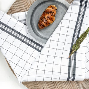 May include: A white and black checkered kitchen towel is folded over a wooden cutting board. A gray ceramic plate with a croissant on it is on top of the towel.