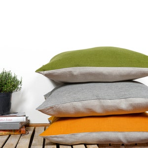 Könnte beinhalten: Drei dekorative Kissen übereinander gestapelt. Das oberste Kissen ist einfarbig grün, das mittlere Kissen hat ein grau-weiß gestreiftes Muster und das unterste Kissen ist einfarbig orange. Jedes Kissen hat einen hellgrauen Leinenrand.
