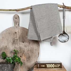 May include: A wooden cutting board with a rustic finish hangs from a branch with a striped linen tea towel and a metal slotted spoon. A small potted plant sits in front of the board. A cookbook titled "1000" is on a wooden cutting board.
