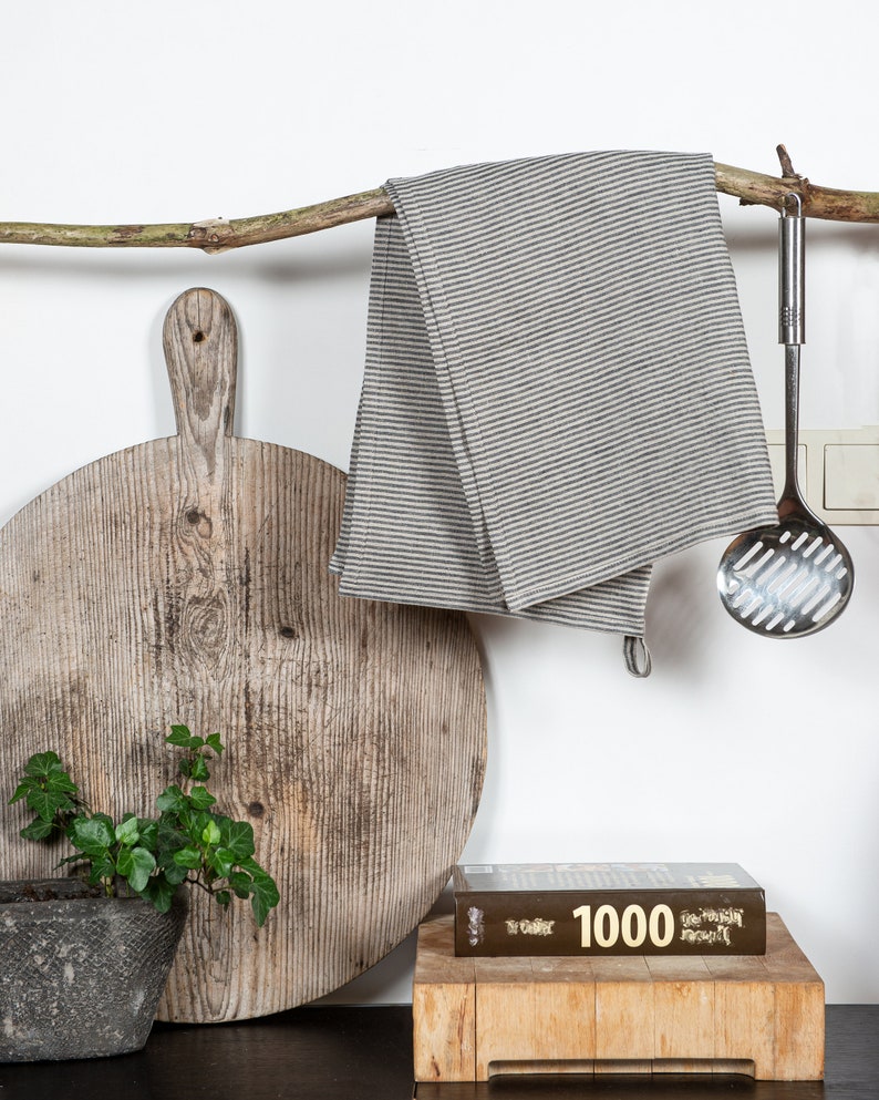 May include: A wooden cutting board with a rustic finish hangs from a branch with a striped linen tea towel and a metal slotted spoon. A small potted plant sits in front of the board. A cookbook titled "1000" is on a wooden cutting board.