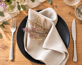 May include: A beige linen napkin folded on a matte black dinner plate, accompanied by a stainless steel fork and knife. A small sprig of dried purple and white flowers rests on the napkin, which is set on a wooden table surface.
