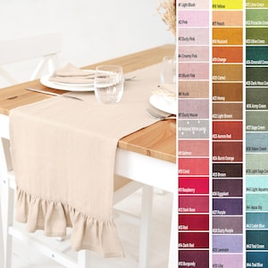 May include: A beige linen table runner with a frilled edge, displayed on a wooden table with place settings. The runner is a neutral colour, complementing the light wood. The image also shows a colour chart with various shades.