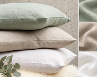 Puede incluir: Una pila de tres cojines cuadrados en verde salvia, beige y blanco, con una textura de tela tejida visible. Los cojines están dispuestos sobre una superficie de piel sintética color crema, acompañados por una pequeña rama de eucalipto.