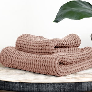 Op de afbeelding: Twee badhanddoeken met een wafelstructuur in een stoffige roze kleur zijn gevouwen en gestapeld op een lichte houten tafel. De handdoeken zijn gemaakt van een zacht, absorberend materiaal.