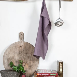 May include: A purple linen kitchen towel hangs on a branch with a metal slotted spoon hanging from the branch. A wooden cutting board, a potted plant, and a stack of cookbooks are on a dark countertop.