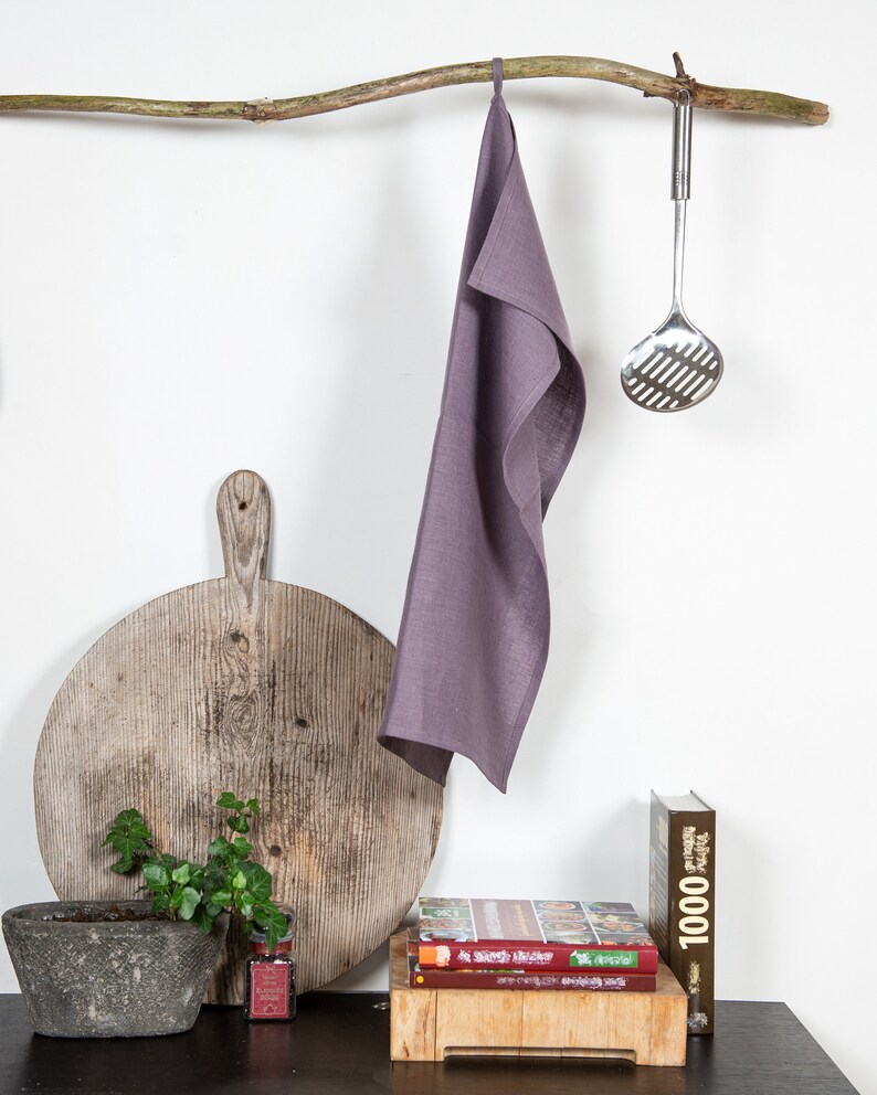 May include: A purple linen kitchen towel hangs on a branch with a metal slotted spoon hanging from the branch. A wooden cutting board, a potted plant, and a stack of cookbooks are on a dark countertop.