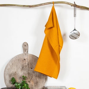 May include: A mustard yellow linen kitchen towel hangs from a branch with a silver slotted spoon hanging from the branch next to it. A wooden cutting board, a potted plant, a cookbook, and bananas are on a table below.
