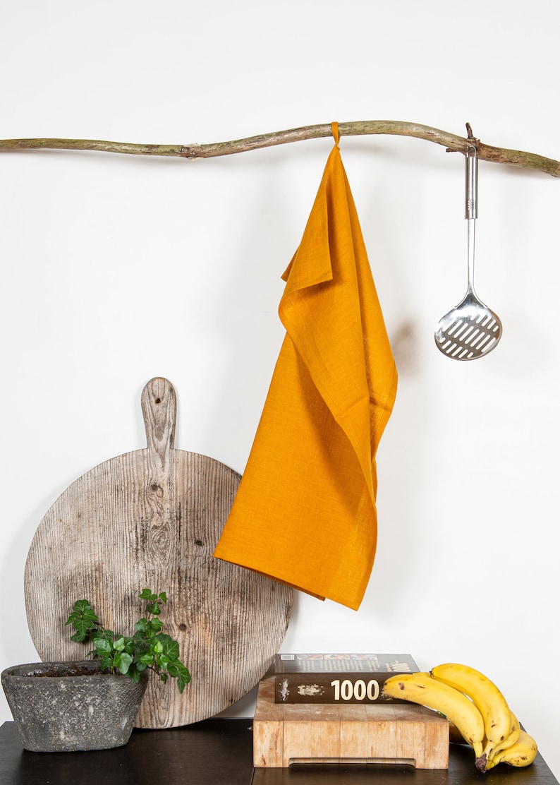 May include: A mustard yellow linen kitchen towel hangs from a branch with a silver slotted spoon hanging from the branch next to it. A wooden cutting board, a potted plant, a cookbook, and bananas are on a table below.