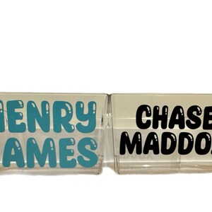 May include: Two clear plastic storage bins with names written on them. The bin on the left says "HENRY JAMES" in teal blue letters. The bin on the right says "CHASE MADDOX" in black letters.