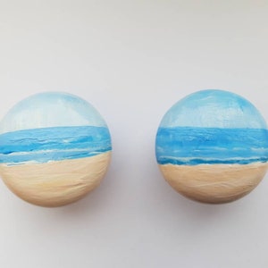 May include: Two round stones painted with a seascape. The top half of each stone is painted a light blue sky with white clouds. The bottom half is painted a blue ocean with white waves and a tan beach.