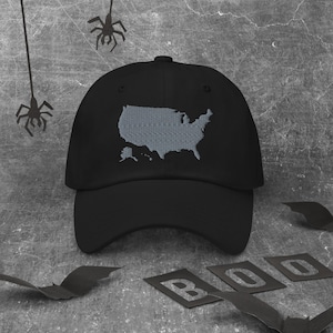 May include: Black baseball cap featuring a grey embroidered outline of the United States. The cap has a classic six-panel design with a curved peak. The background includes Halloween decorations such as bats and spiders.